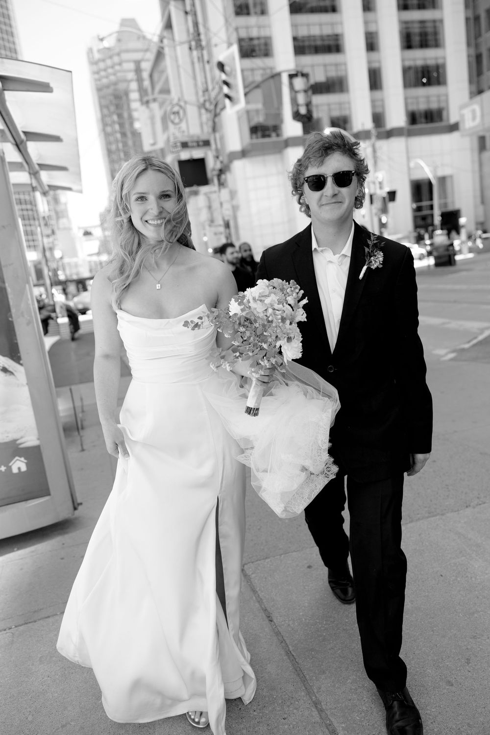 Bride and groom walking on a city street, bride in white gown holding bouquet, groom in black suit with sunglasses.