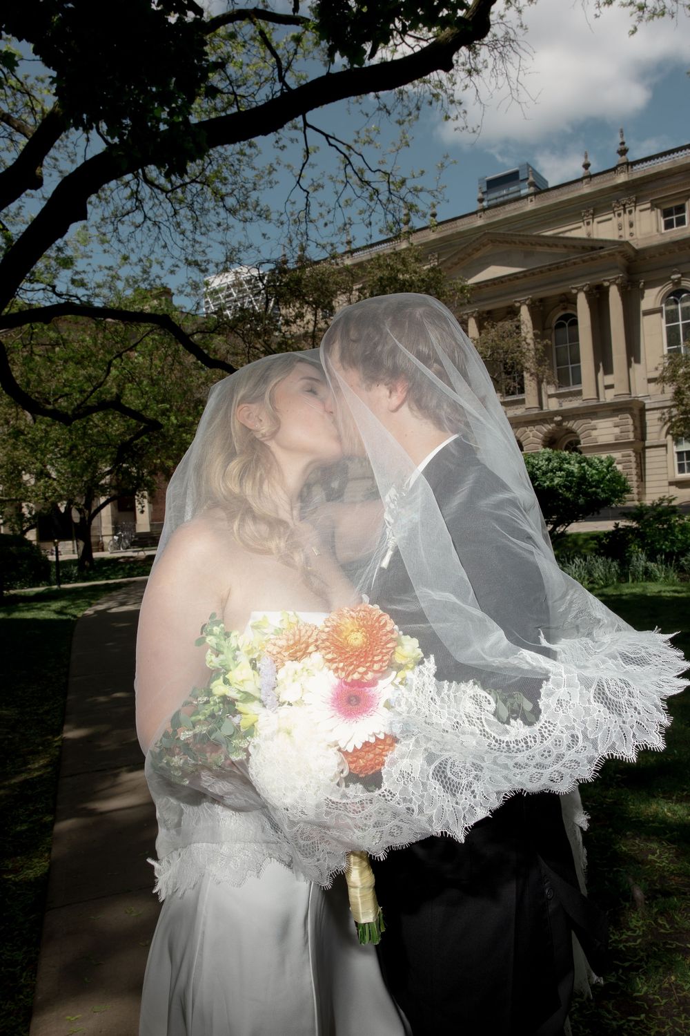 Bride and groom kissing under a lace veil outdoors, bride holding colorful bouquet near a historic building.