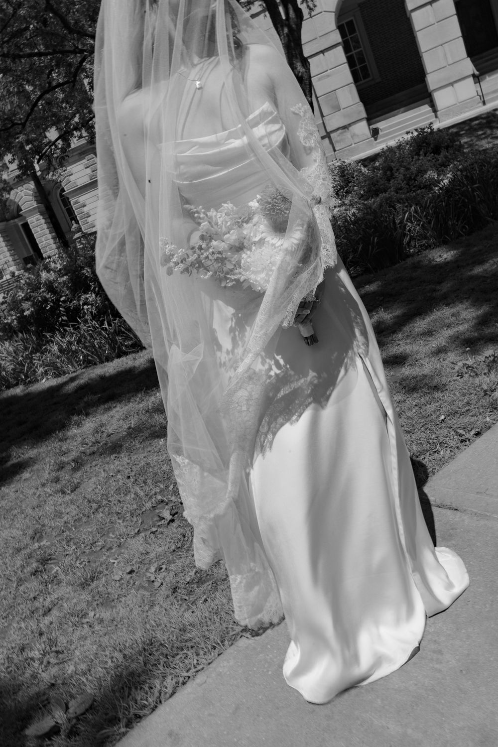 Black and white photo of a bride in a white gown and veil holding a bouquet outdoors in sunlight.