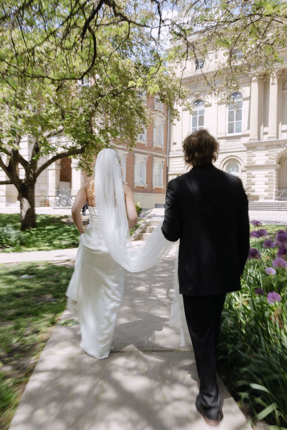 Bride and groom walking hand in hand along a garden path toward a classic brick building in spring.