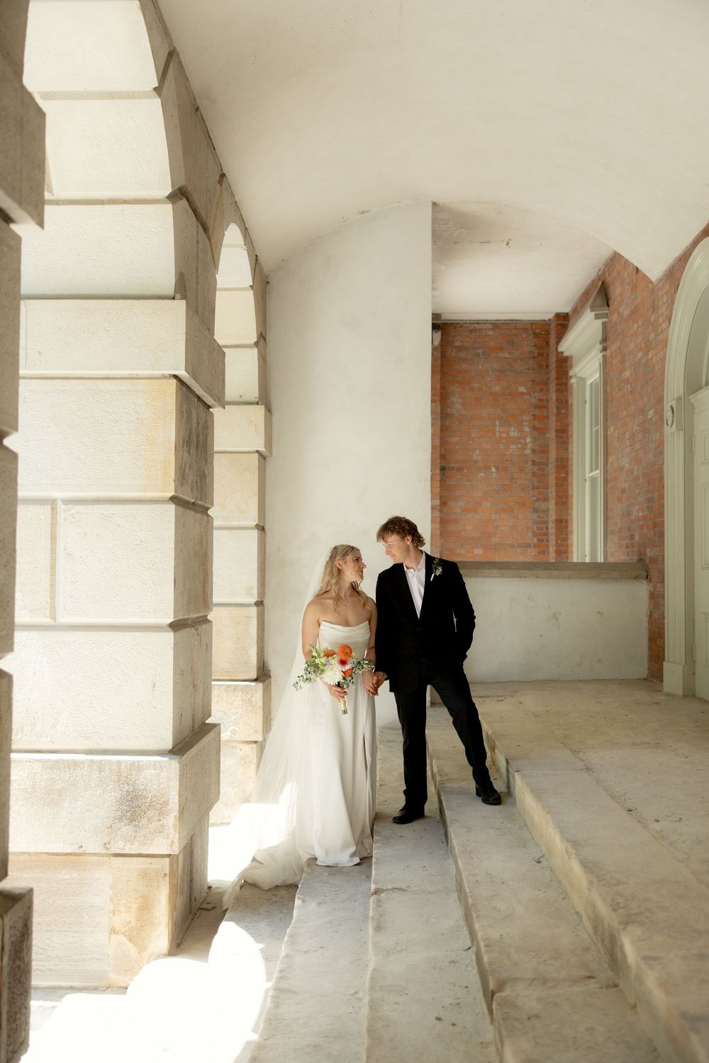 Bride and groom standing together in a sunlit stone archway corridor of a historic building.
