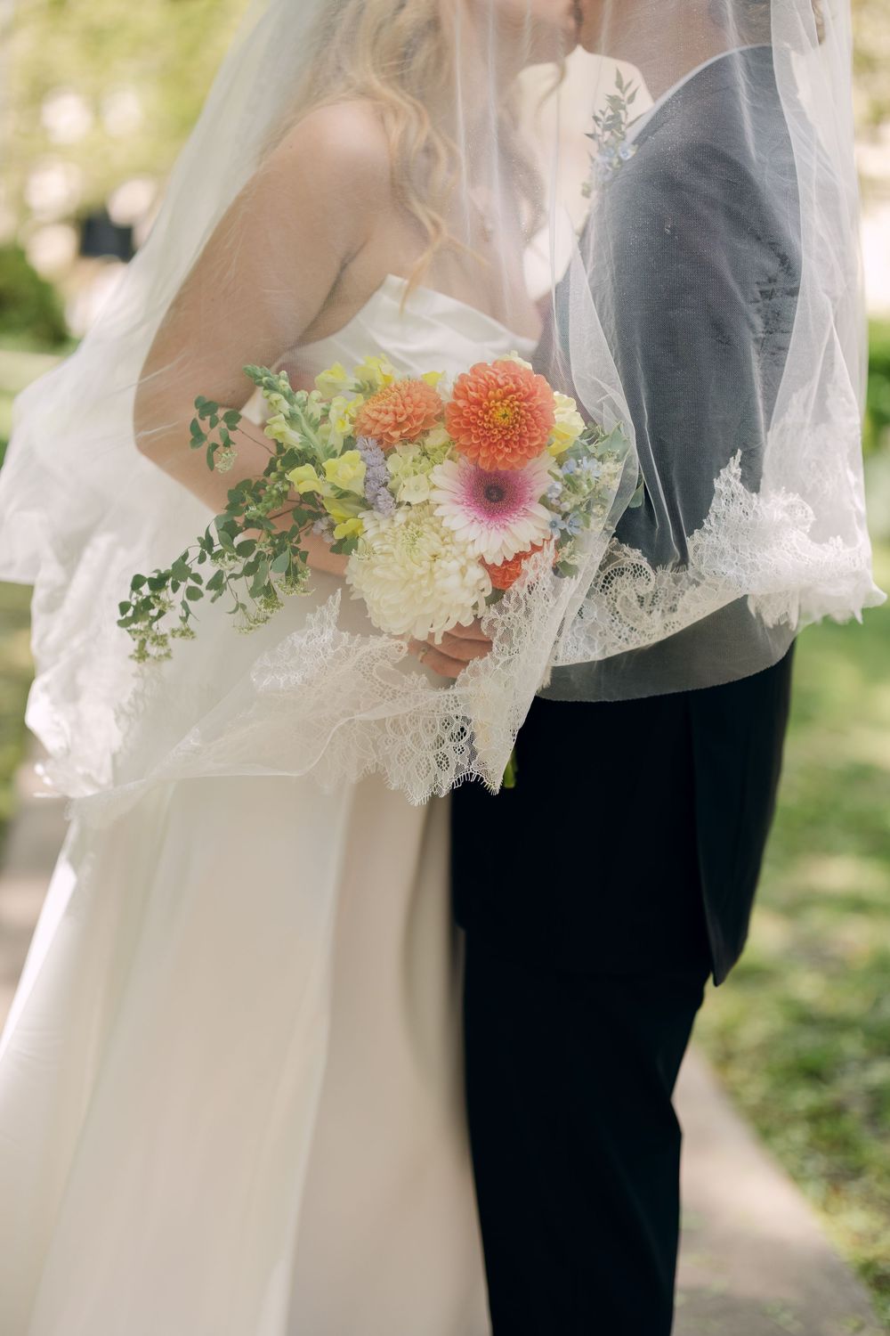 Bride and groom share a romantic moment under a lace wedding veil, bride holding a colorful bouquet outdoors.