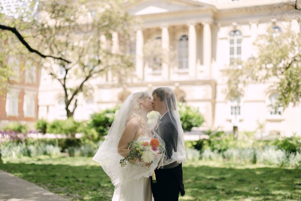 Bride and groom sharing a kiss under a veil outside a historic building surrounded by spring greenery.