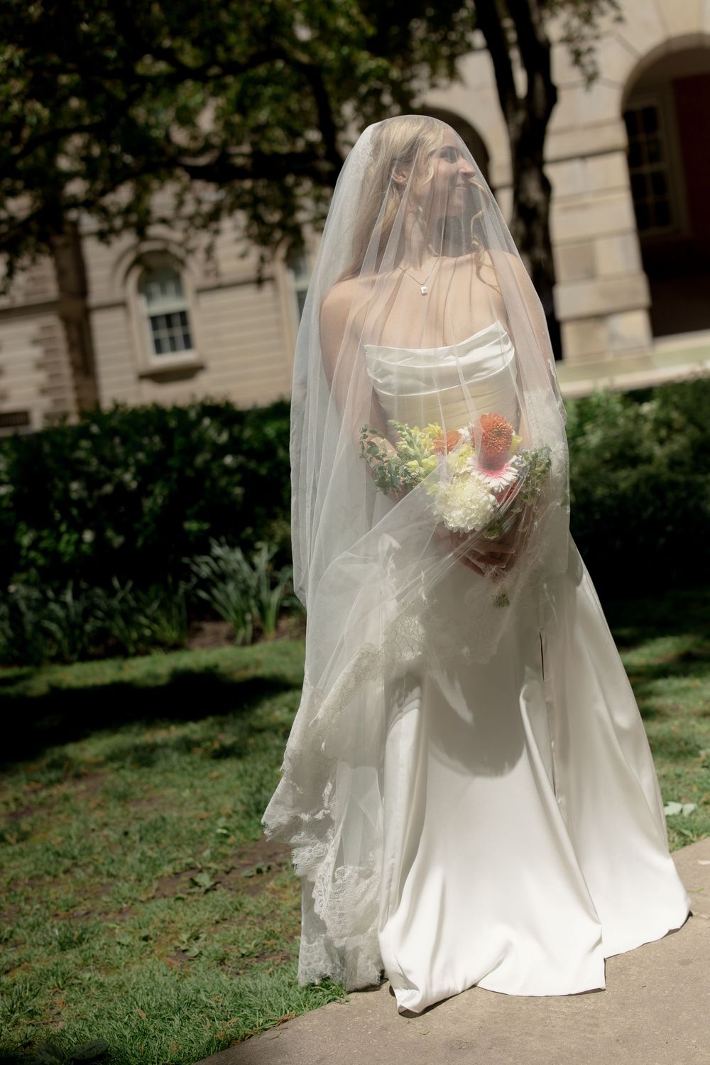 Elegant bride in a strapless white gown and long lace veil holds a bouquet while walking in a garden.