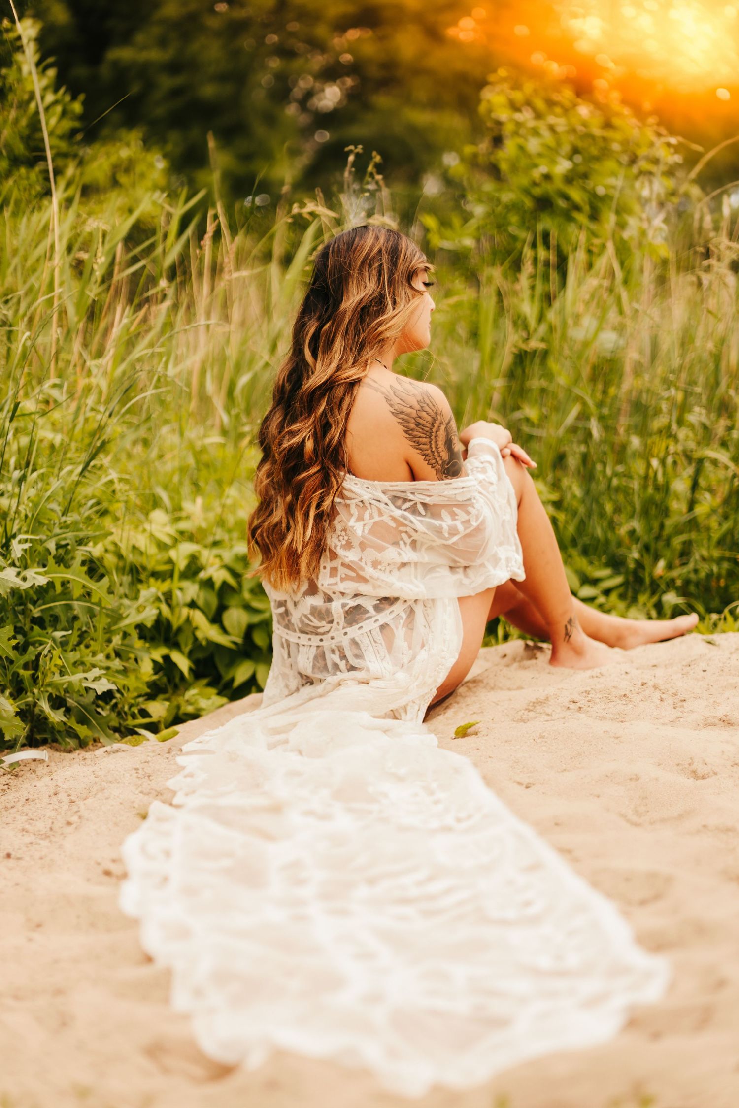 A delicate white lace dress flows across sandy ground as sunlight streams through tall meadow grass at golden hour.