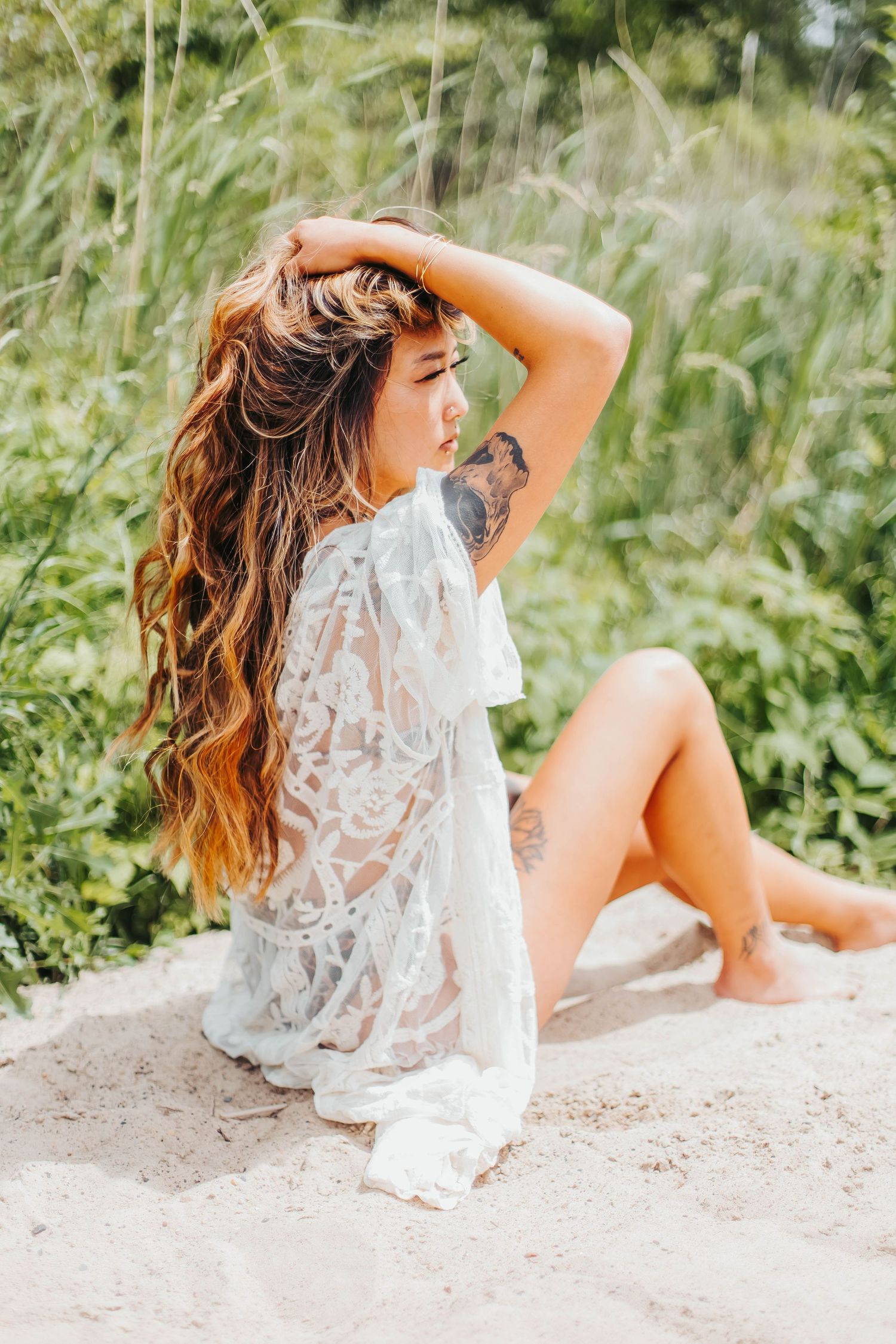 Woman in white lace dress sits on sandy ground surrounded by tall summer grass and greenery in sunlight.