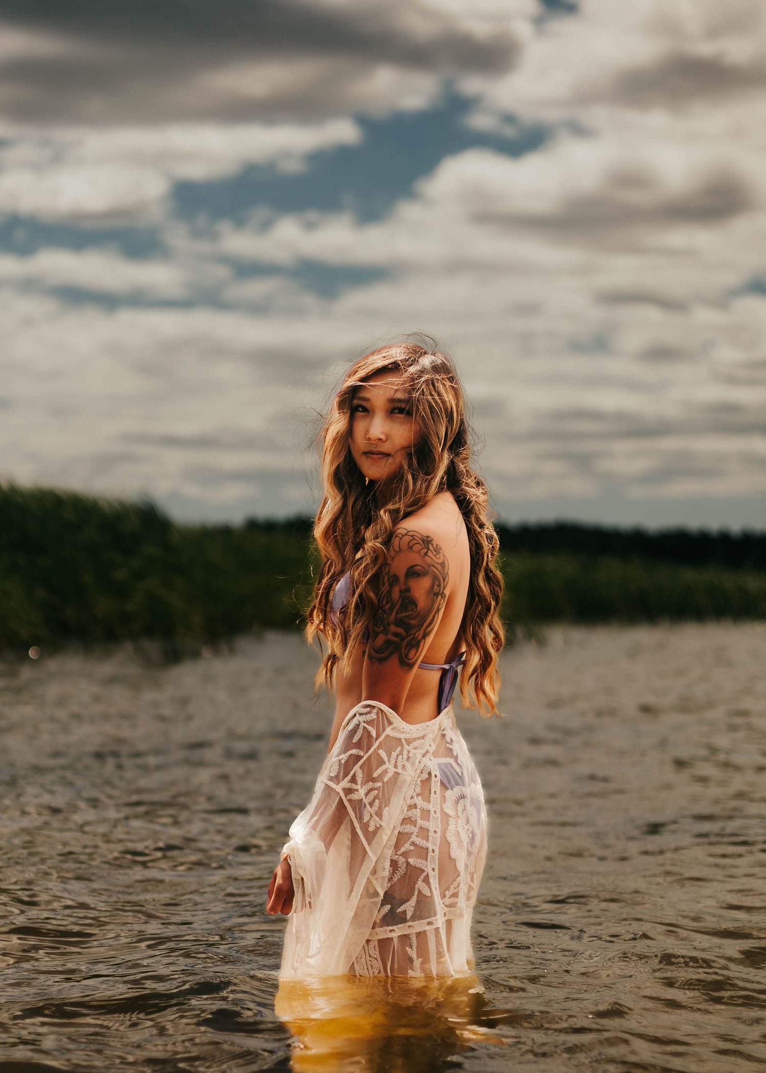 Figure in a delicate lace dress stands in water under dramatic cloudy skies near a forested shore.