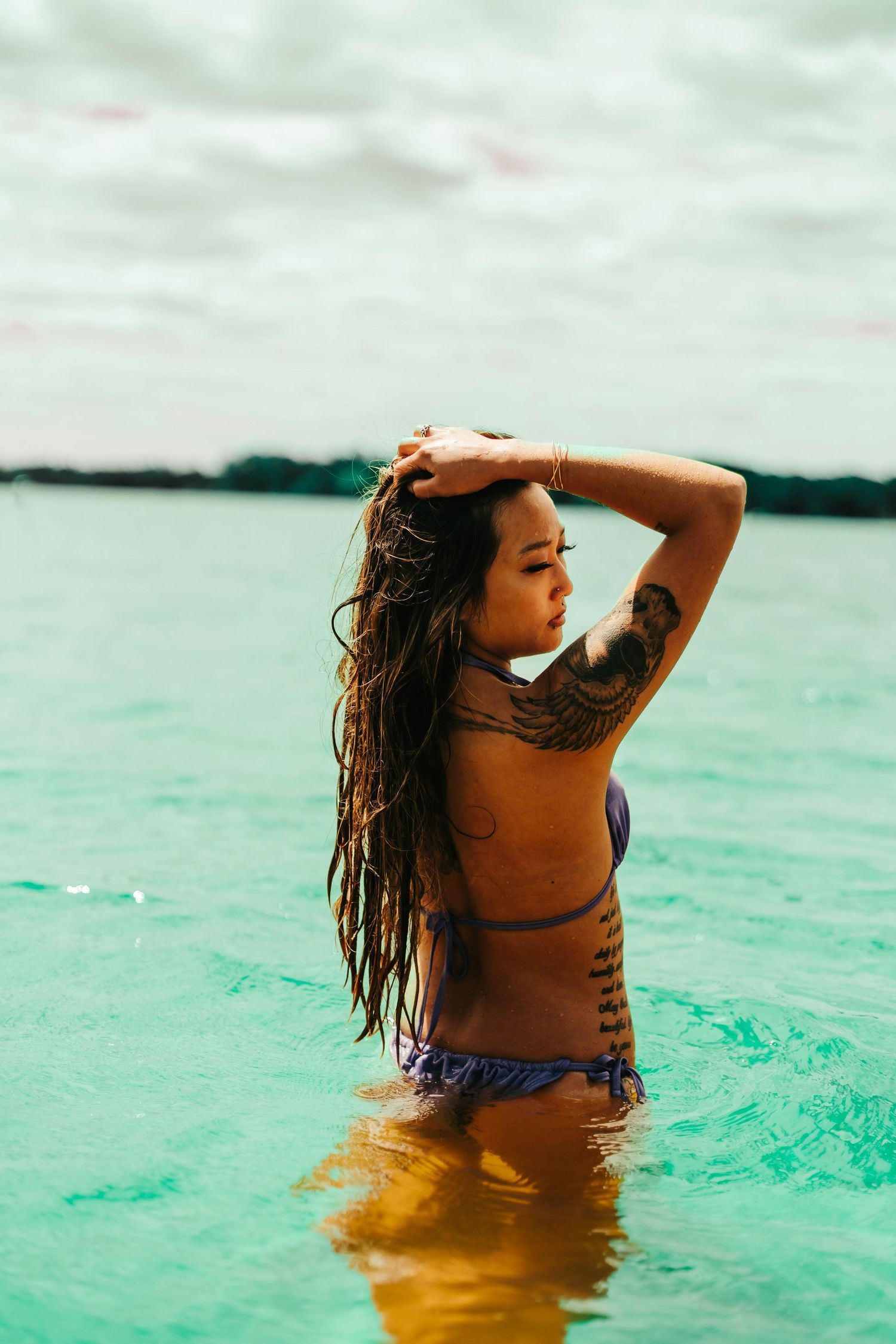 Woman with long dark hair standing in turquoise tropical waters on an overcast day.