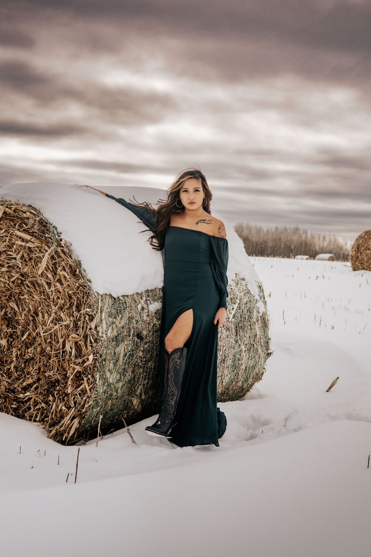A person in a dark dress poses gracefully against rustic hay bales in a snowy winter landscape at dusk.