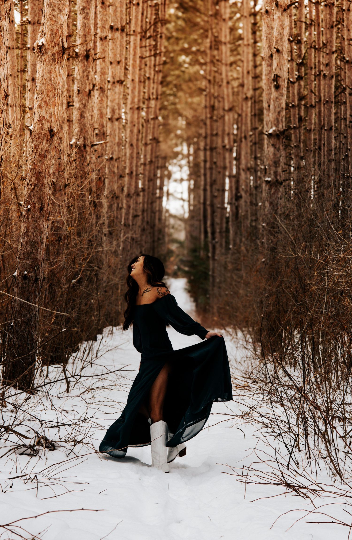 A person in a black dress twirls gracefully in a snowy winter forest with tall bare trees in warm light.