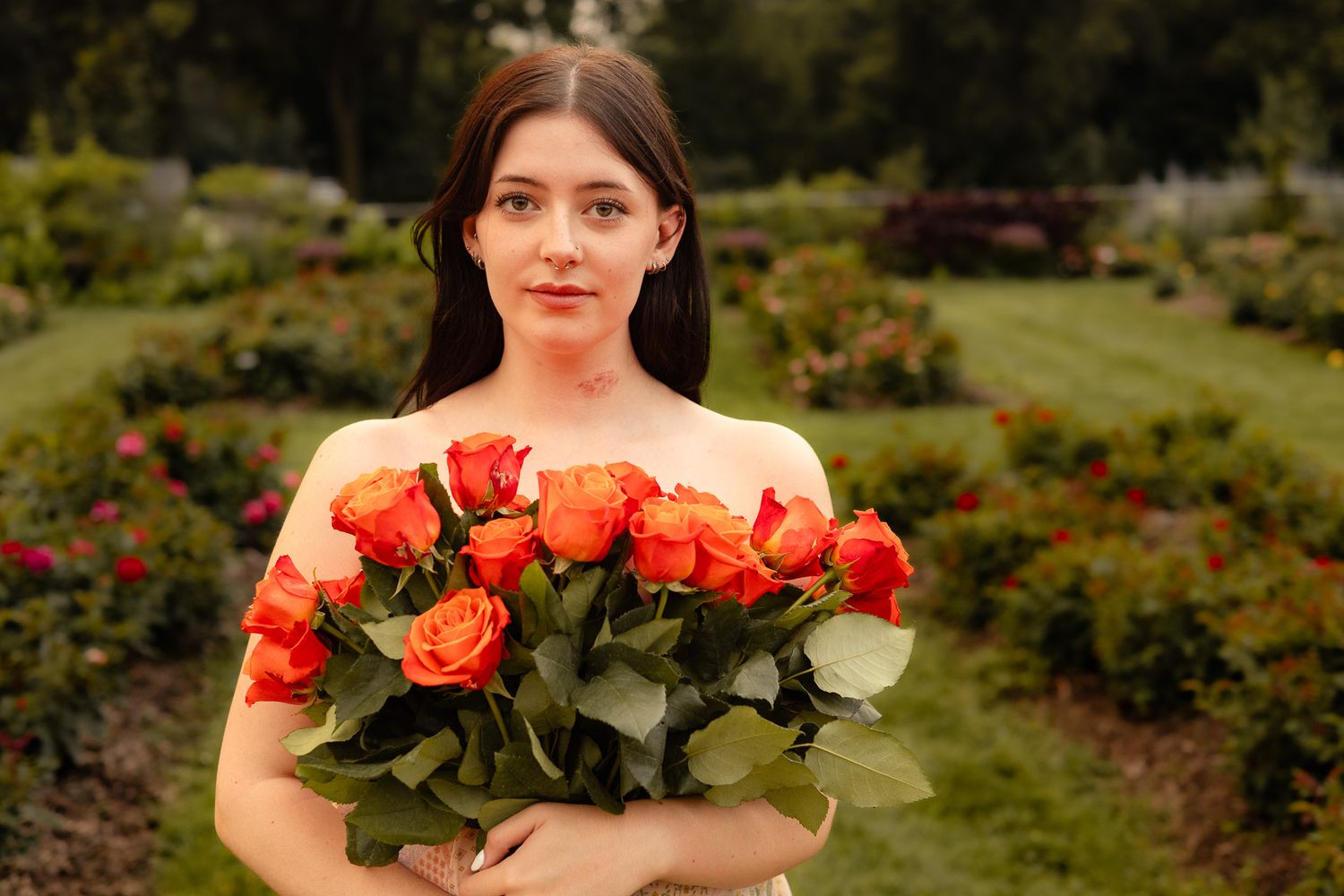 Young woman in garden holding large bouquet of orange roses, surrounded by lush greenery and colorful flowers.
