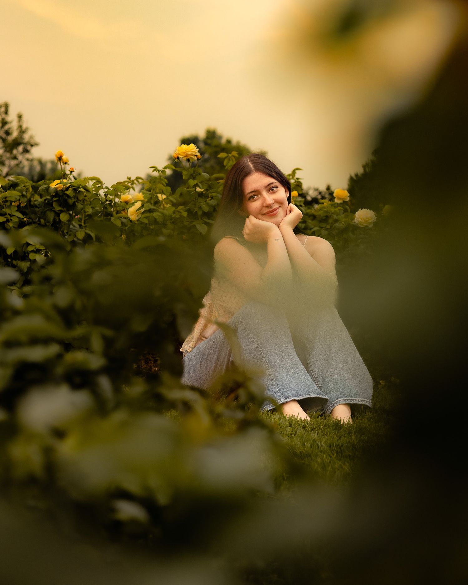 Young woman sitting peacefully among blooming yellow roses in a lush garden at golden hour, smiling softly.
