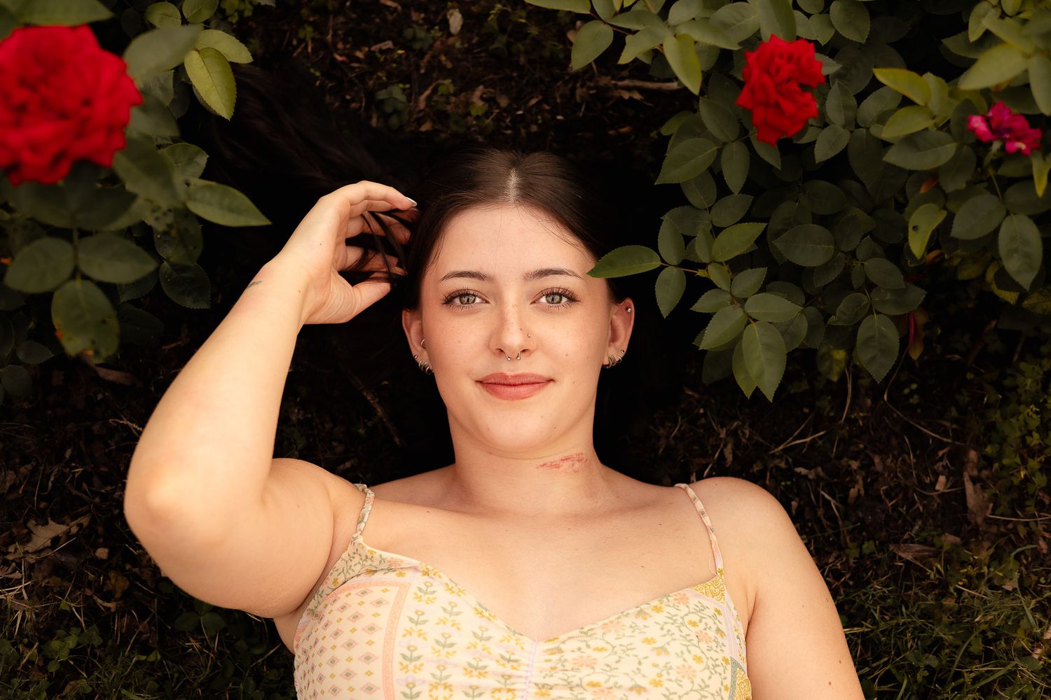 Young woman lying on grass surrounded by red roses and green leaves, smiling up at camera in a floral dress.