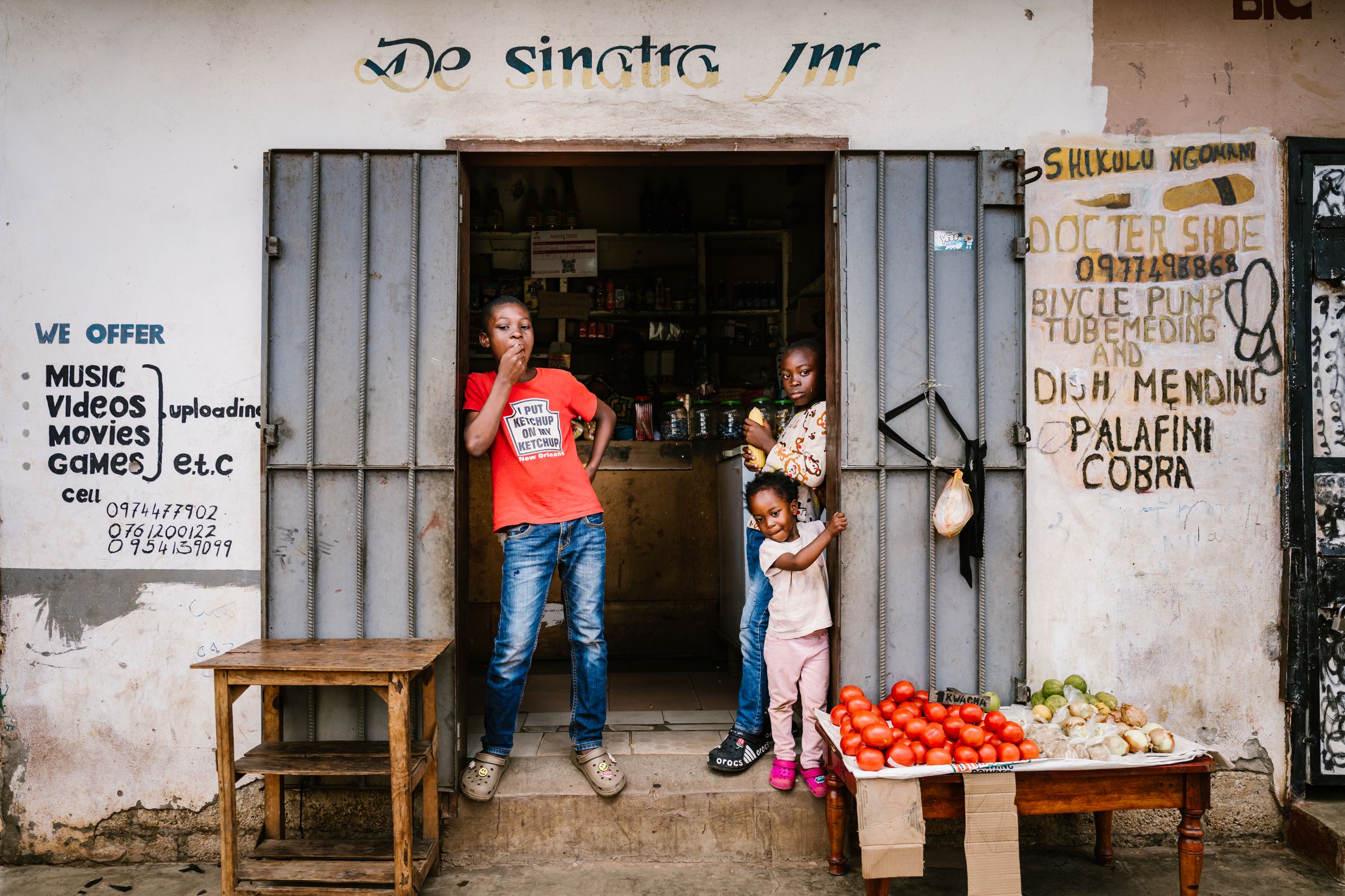 A family stands outside a small African shop with handwritten signs, fresh tomatoes and vegetables displayed on a wooden table.