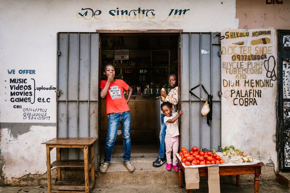 A family stands outside a small African shop with handwritten signs, fresh tomatoes and vegetables displayed on a wooden table.