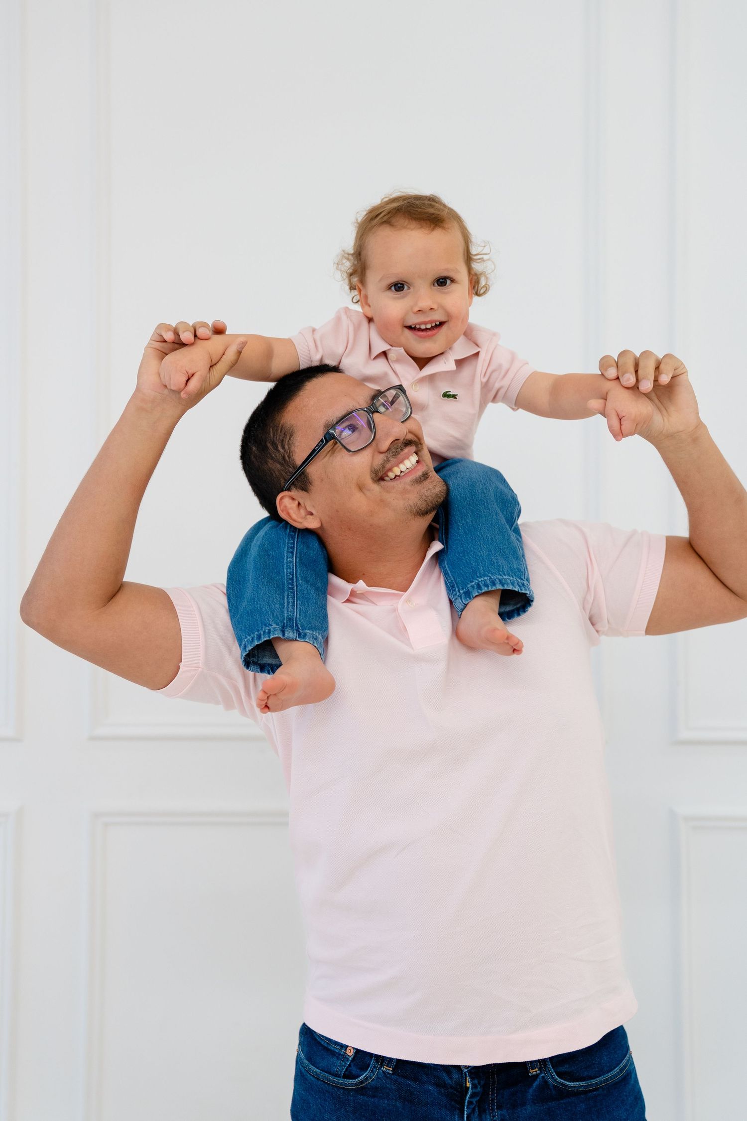 Happy father carrying smiling toddler on shoulders, both wearing pink shirts, posing against white wall.