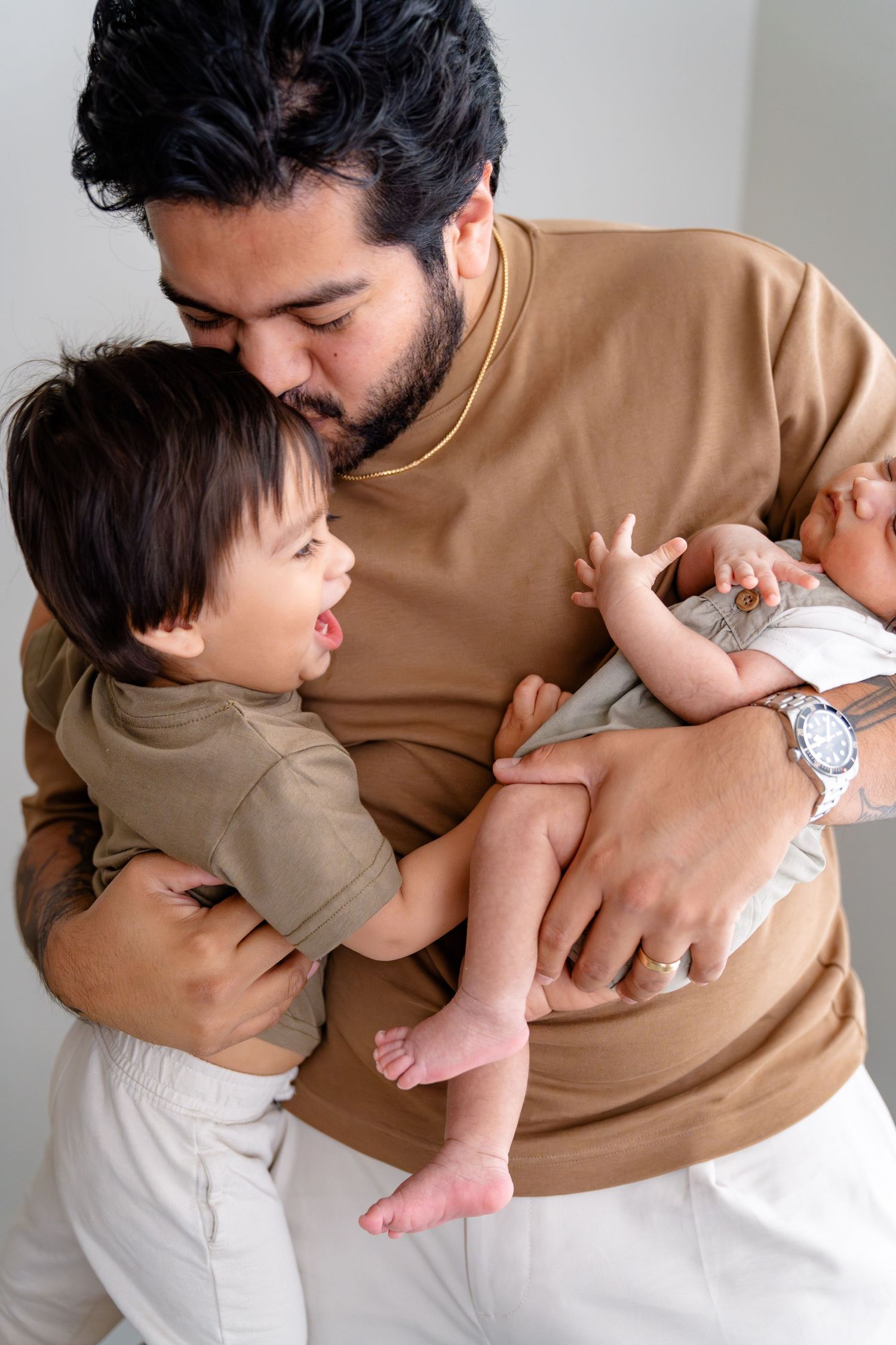 Middle-aged father with gray hair cradling sleeping newborn baby wrapped in beige knit blanket near bright window.