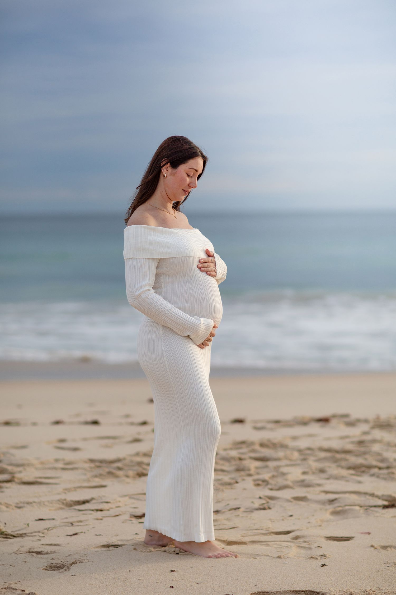 Mum to be in white dress admiring her baby bump on the beach during maternity session at golden hour in perth
