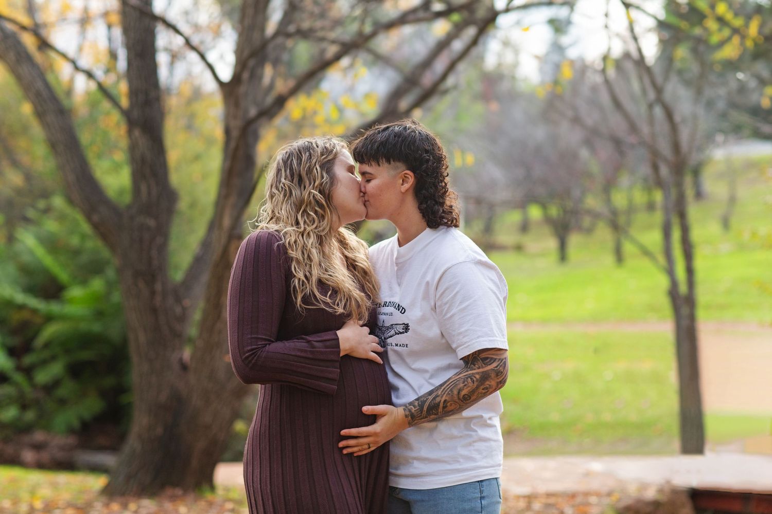 Couple sharing a kiss while holding baby bump surrounded by trees during autumn in Perth