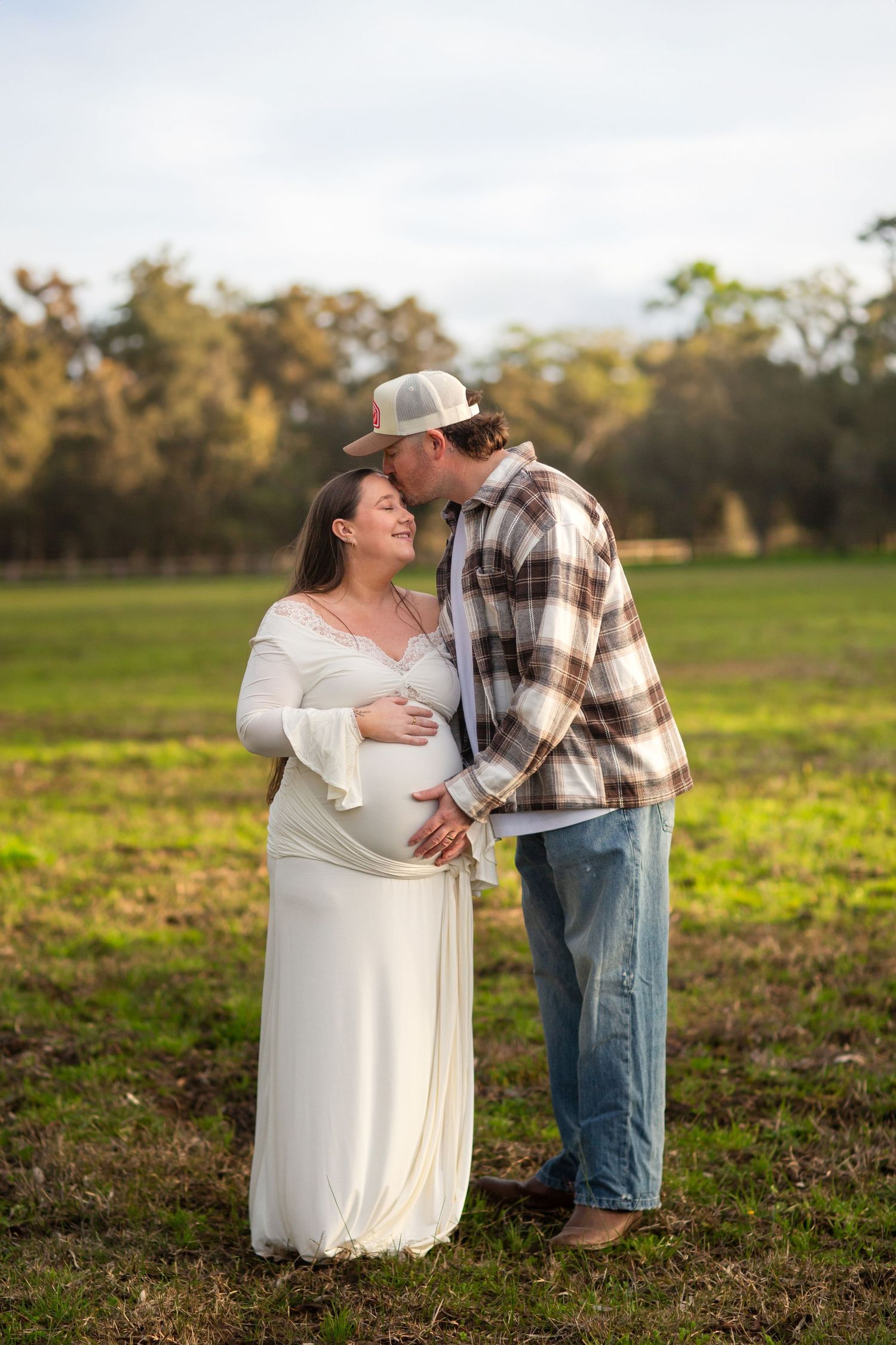 Partner kissing mum to be while holding baby bump during golden hour in a paddock during maternity session in Perth