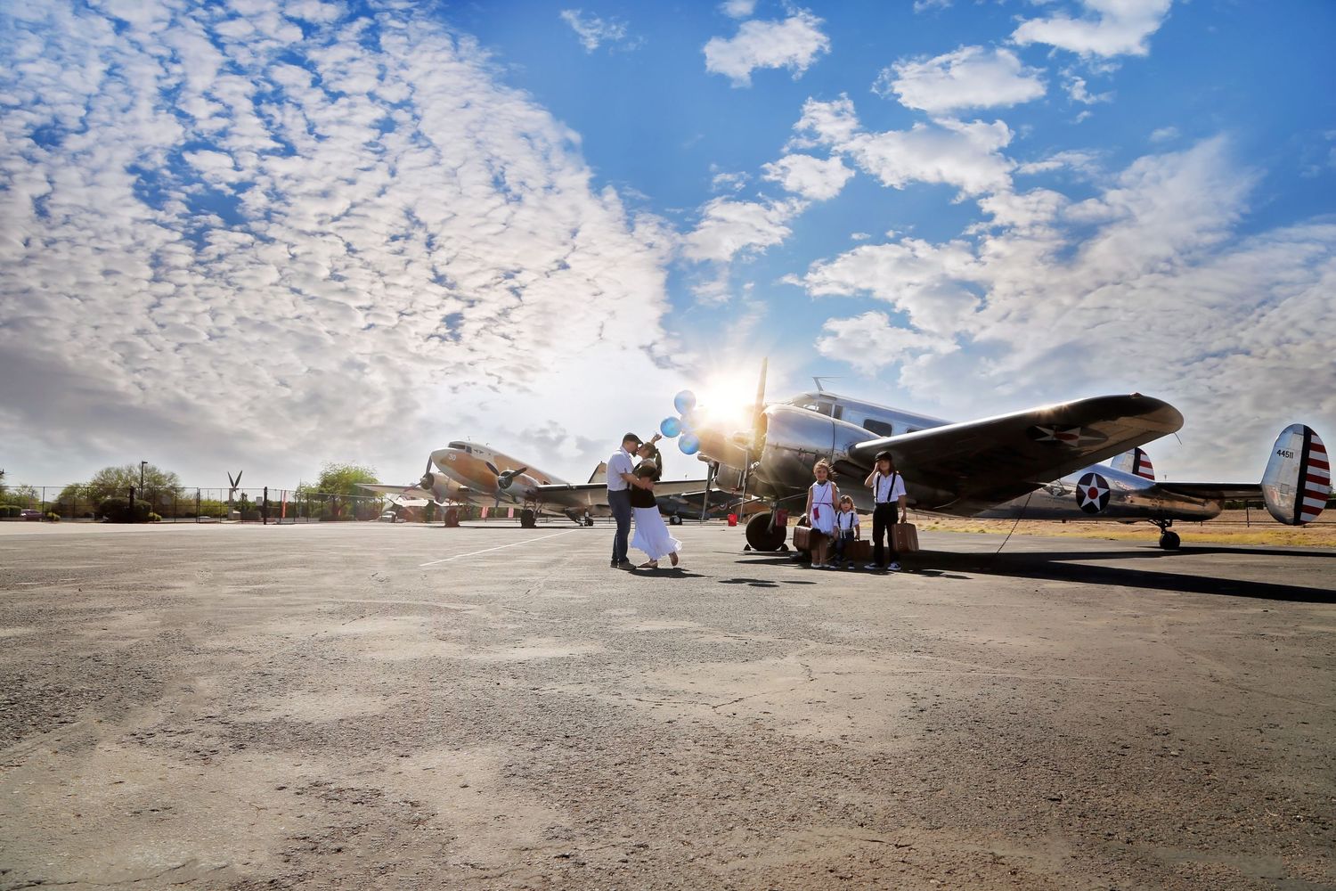 Wedding couple stands near vintage airplanes on an airfield under a bright blue sky with dramatic clouds and sunburst.
