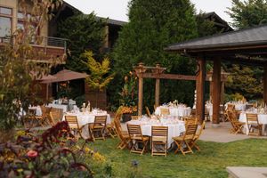 Elegant outdoor wedding reception with wooden chairs and white-draped tables on a lush garden patio beside a rustic pergola.