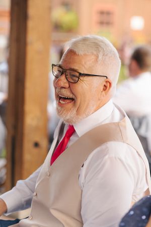 Elderly man with white hair and beard, wearing glasses and red tie, laughing joyfully at a formal event.