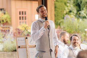 Man in gray vest giving a speech with microphone at an outdoor wedding reception.