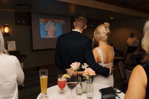 Bride and groom watch a projected photo at their wedding reception, with floral centerpiece and drinks on table.