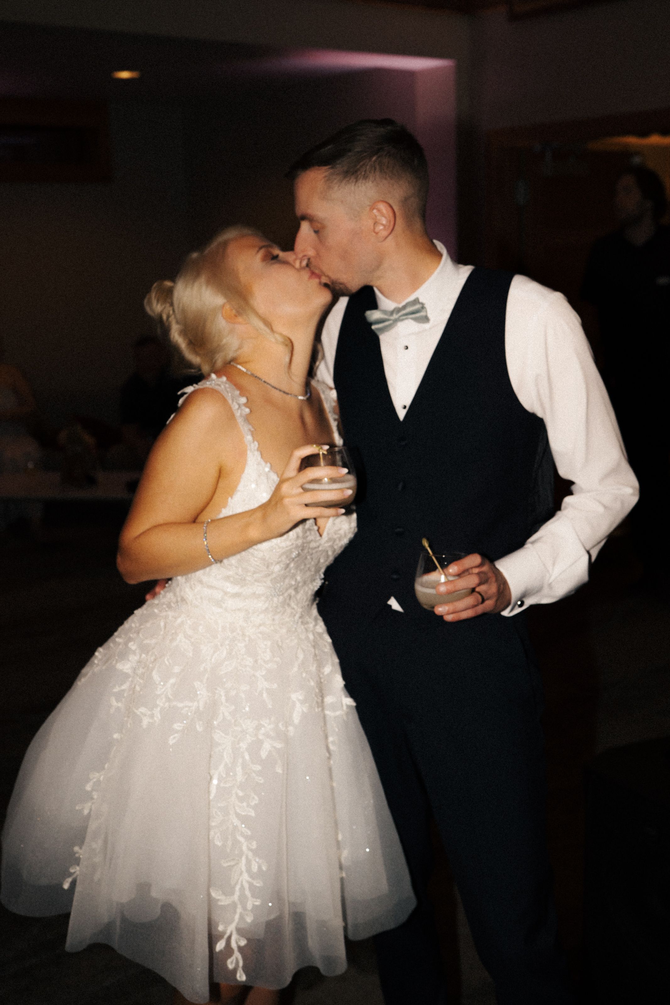 Bride in short white lace dress and groom in black vest with mint bow tie share a kiss while holding drinks.