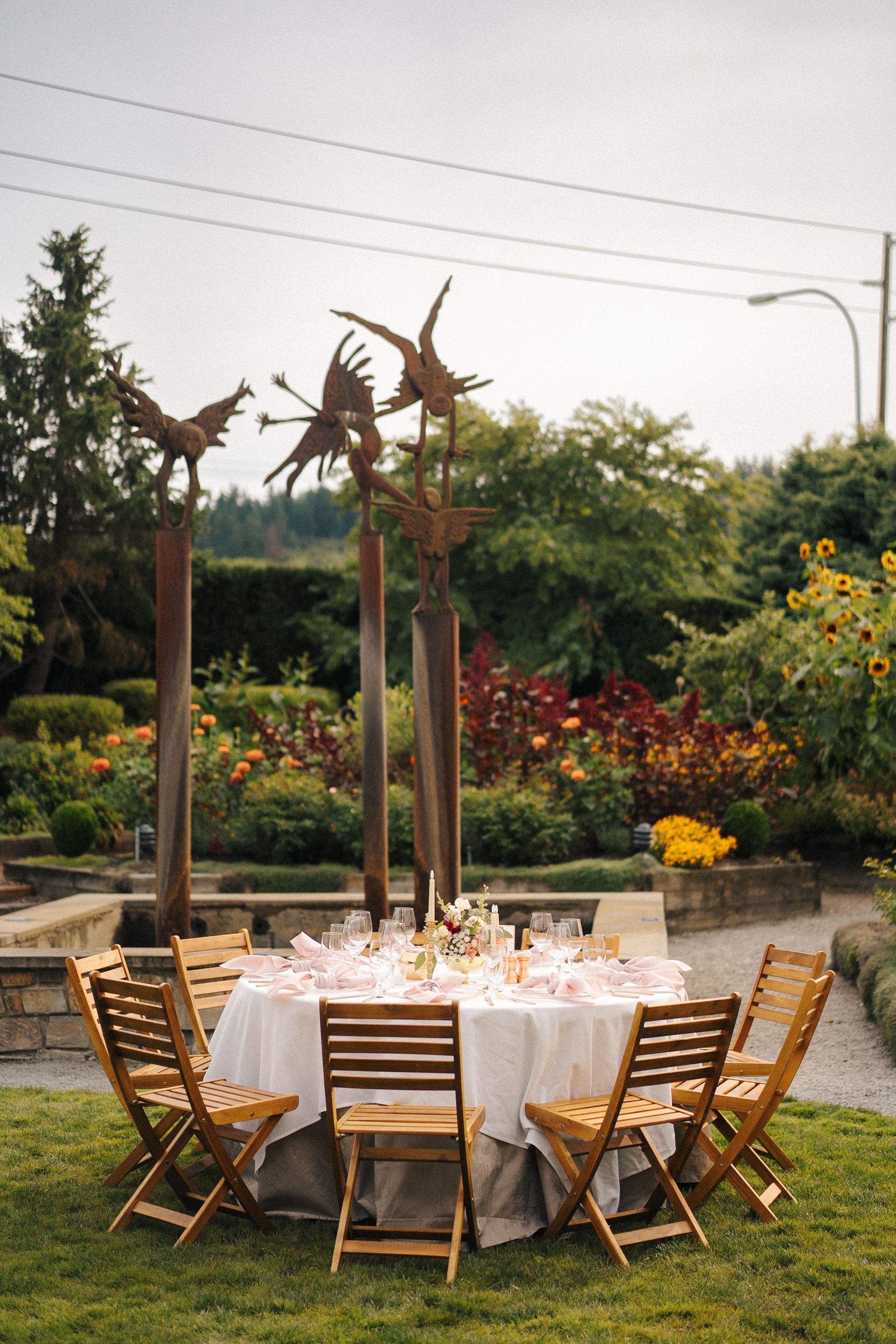 Outdoor garden wedding reception table with wooden chairs, white tablecloth, and rustic metal bird sculptures amid colorful flowers.