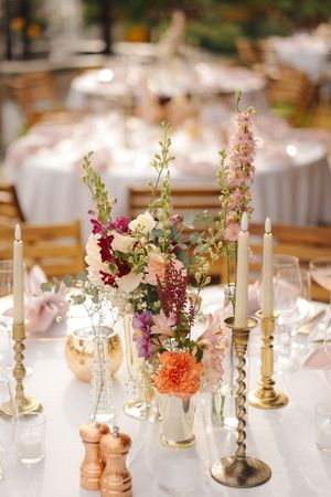Elegant wedding table centerpiece with colorful flowers in gold vases, taper candles, and glassware on a white tablecloth.