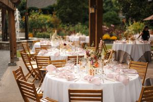 Elegant outdoor wedding reception with round tables, white linens, pink accents, floral centerpieces, and wooden chairs under a pergola.