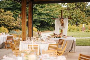 Elegant outdoor wedding reception setup with wooden chairs, white tablecloths, floral arch, and lush garden backdrop.