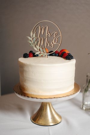 White wedding cake with Mr & Mrs topper, fresh berries, on gold stand.