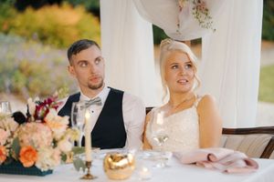Bride and groom seated at wedding reception table with floral centerpiece and champagne glasses.