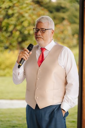 Older man with gray hair and glasses giving a speech with microphone, wearing vest and red tie outdoors.