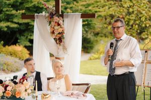 Man giving speech at outdoor wedding reception while bride and groom sit at decorated table with floral arrangements.
