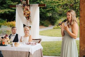 Bridesmaid giving a heartfelt speech at an outdoor wedding reception while the bride and groom listen at their table.