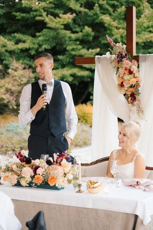 Groom giving a speech at wedding reception with bride seated at floral-decorated head table outdoors.