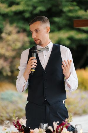 A groom in a black vest and bow tie speaks into a microphone while gesturing during a wedding reception speech outdoors.