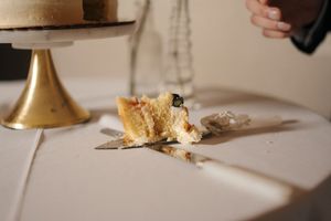 A fallen wedding cake slice lies on a white tablecloth beside a gold cake stand, with a hand reaching in the background.