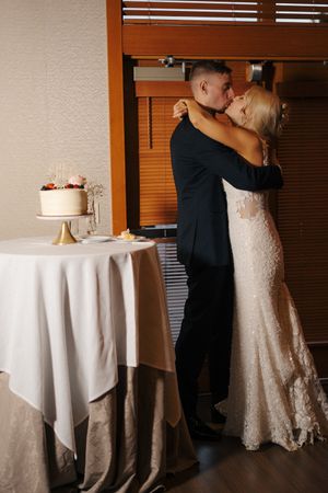 Bride and groom sharing a kiss beside a white wedding cake on a draped table in an elegant indoor venue.