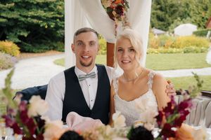 A smiling bride and groom in wedding attire seated at a floral reception table outdoors.