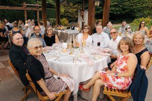 Guests smiling at a round table during an elegant outdoor wedding reception with white tablecloths and floral centerpieces.