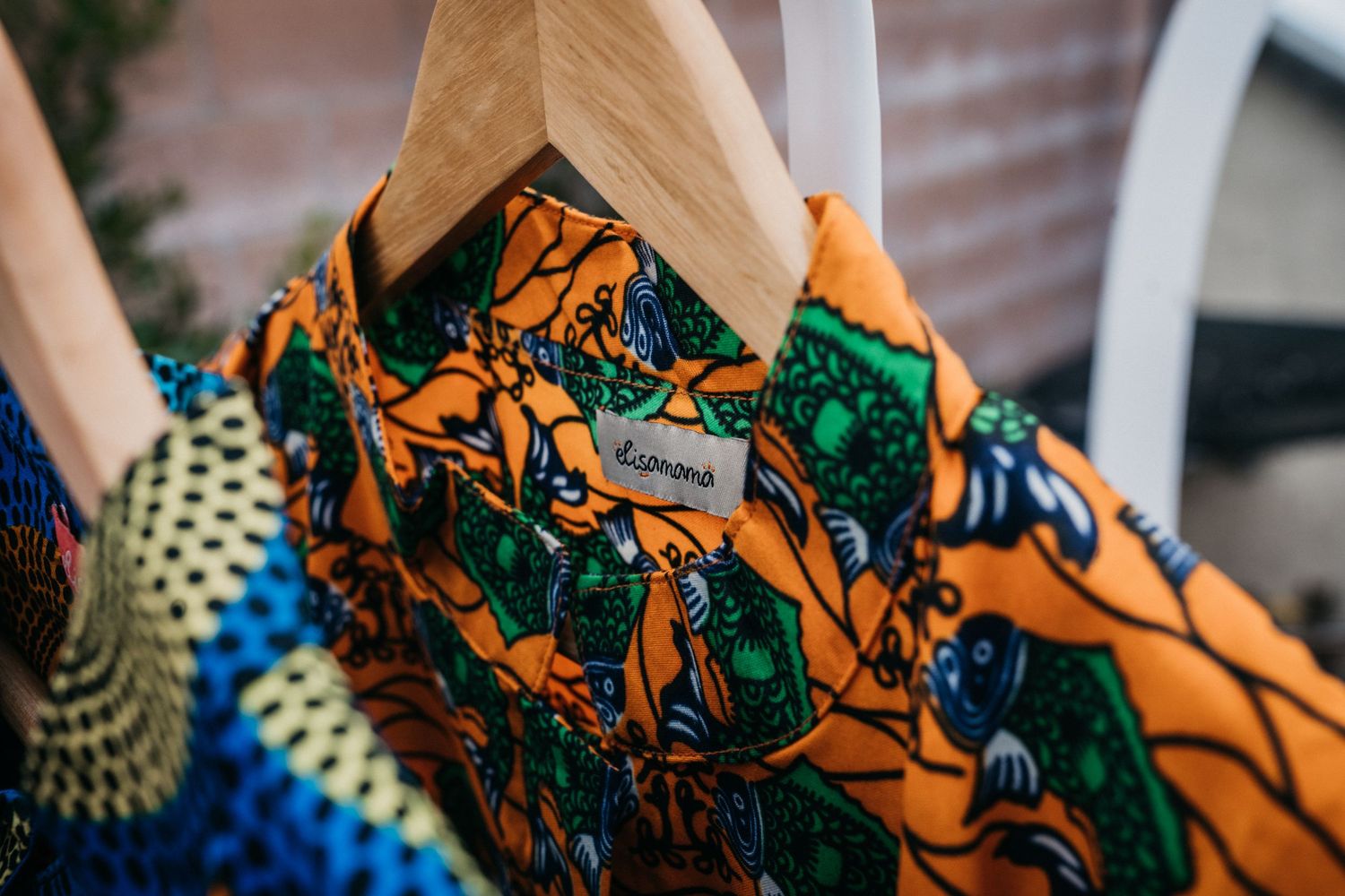 Colorful African print shirt on a wooden hanger displaying vibrant orange, blue, and green patterns with a brand label.