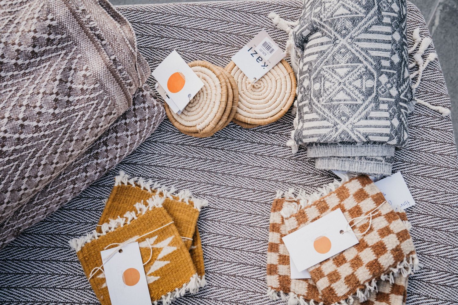Flat lay of handwoven textiles, wicker baskets, and checkered fabric coasters with price tags on a gray woven surface.