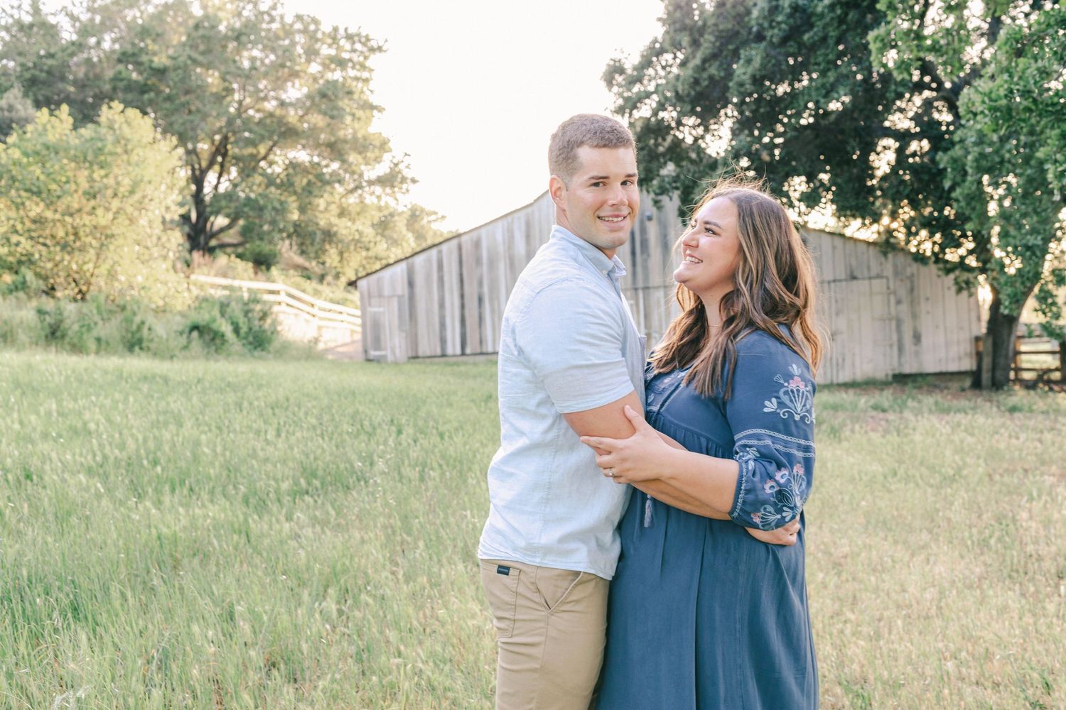 Couple embracing and smiling in a sunny green field with a rustic barn in the background.