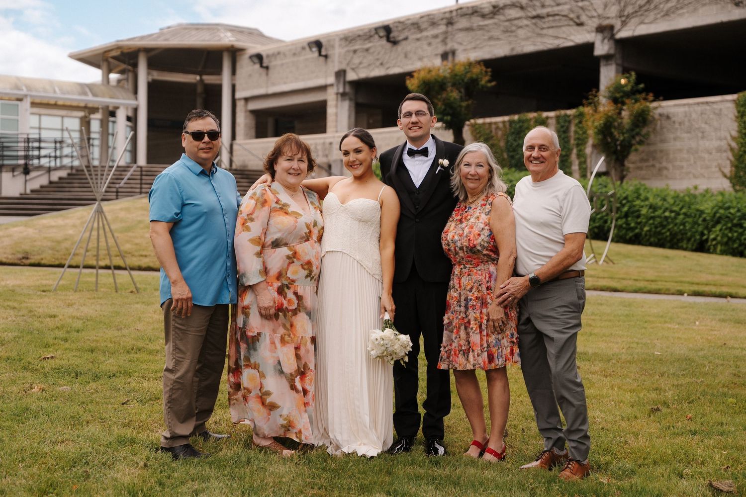 Wedding couple poses with four family members outdoors on grass in front of a modern building on a sunny day.