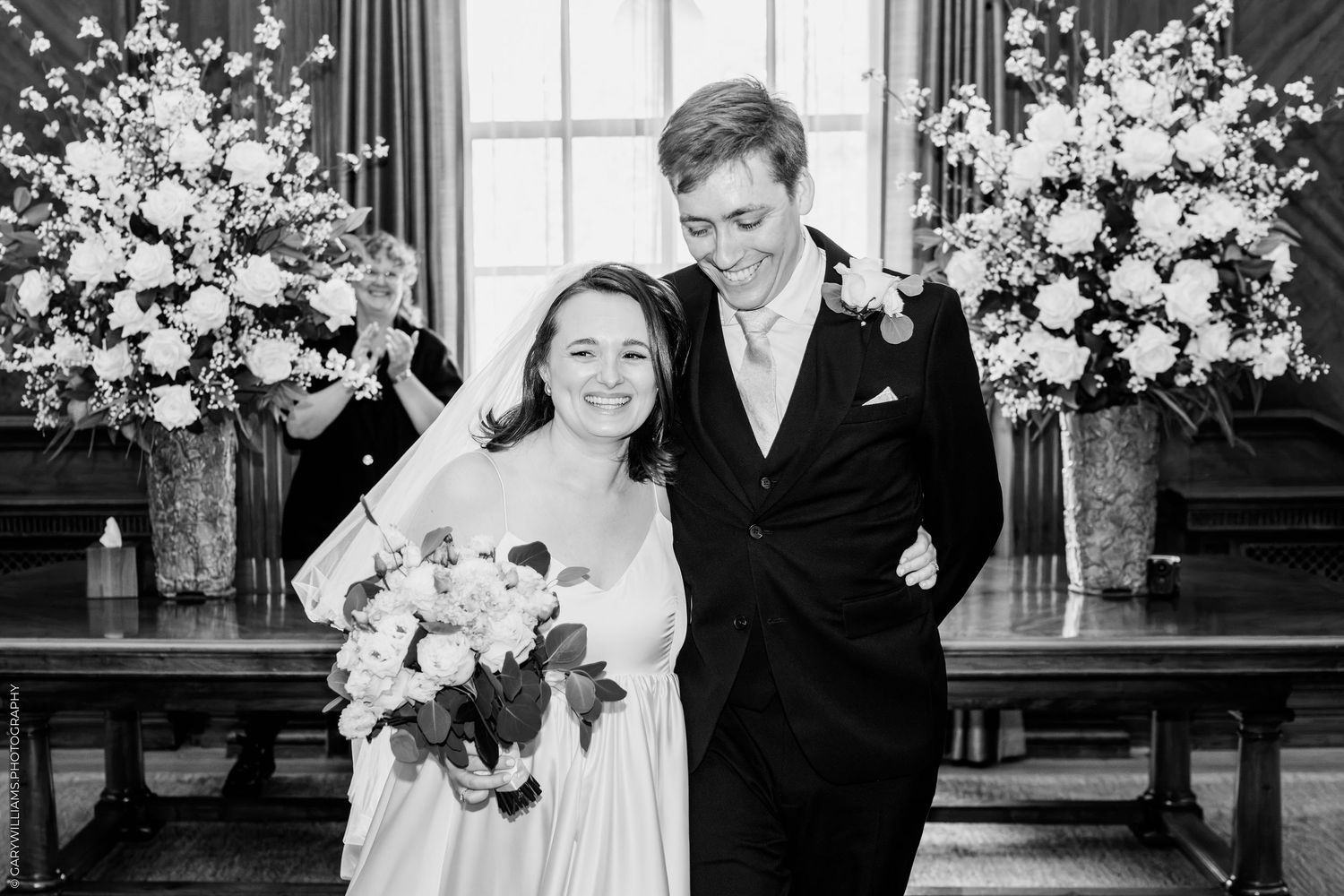 Black and white photo of a smiling bride holding bouquet and groom in suit at wedding ceremony with floral arrangements.